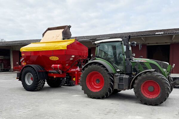 Du siehst einen grünen Traktor mit rotem Düngerstreuer beim Beladen auf einem landwirtschaftlichen Hof.