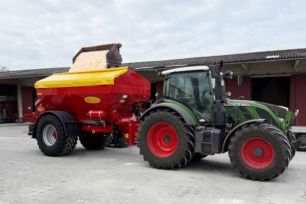 Du siehst einen grünen Traktor mit rotem Düngerstreuer beim Beladen auf einem landwirtschaftlichen Hof.