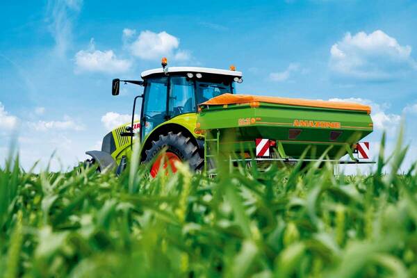 Du siehst einen grünen Traktor mit Amazone-Düngerstreuer bei sonnigem Wetter auf einem Feld.