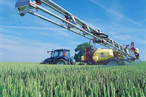 Du siehst einen Traktor mit Pflanzenschutzspritze bei der Feldarbeit in einem grünen Getreidefeld.