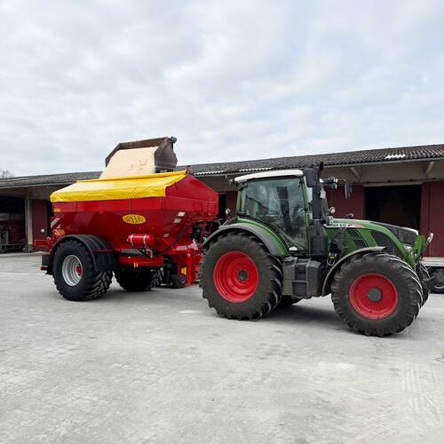 Du siehst einen grünen Traktor mit rotem Düngerstreuer beim Beladen auf einem landwirtschaftlichen Hof.