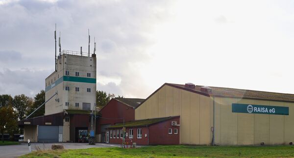Du siehst eine Lagerhalle mit einem grünen RAISA eG-Schild und einen Turm zur Lagerung von Getreide