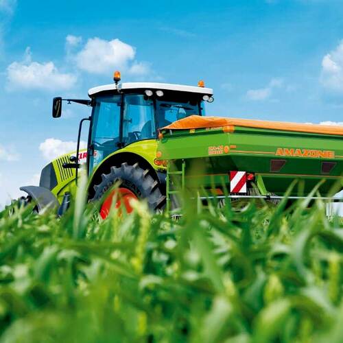 Du siehst einen grünen Traktor mit Amazone-Düngerstreuer bei sonnigem Wetter auf einem Feld.
