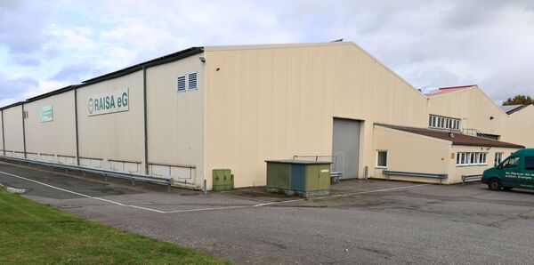 Du siehst eine Lagerhalle mit einer grünen Aufschrift RAISA eG sowie eine Parkfläche davor