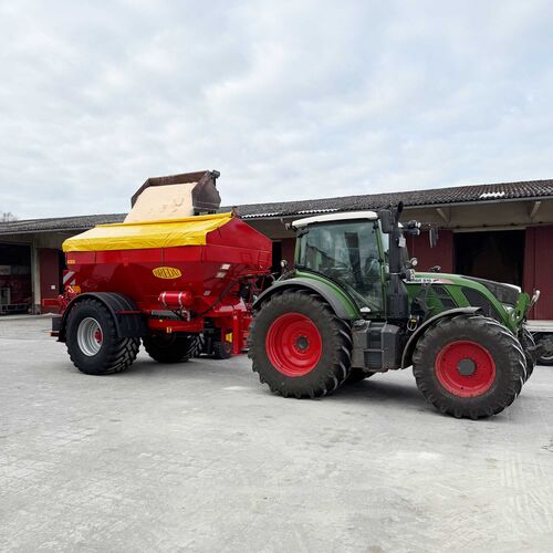 Du siehst einen grünen Traktor mit rotem Düngerstreuer beim Beladen auf einem landwirtschaftlichen Hof.