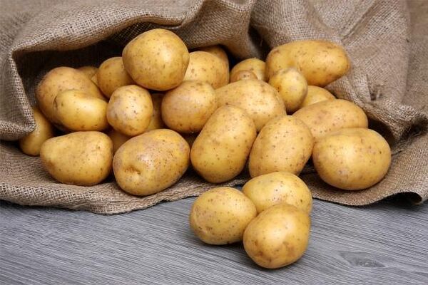 Du siehst frisch geerntete, saubere Kartoffeln, die aus einem Jutesack auf einen Holztisch rollen.